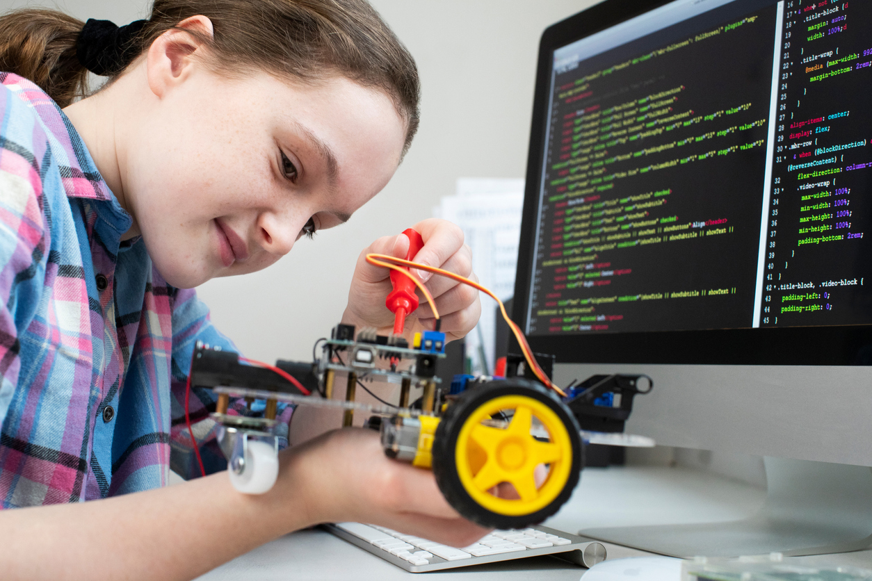 Young person working on a robotics project, using a screwdriver on a small vehicle with yellow wheels. A computer screen displaying lines of code is visible in the background.