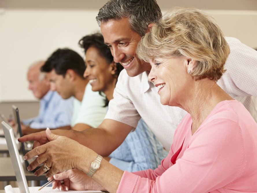 Various people in a computer class with a medium complexion man helping a light complexion woman