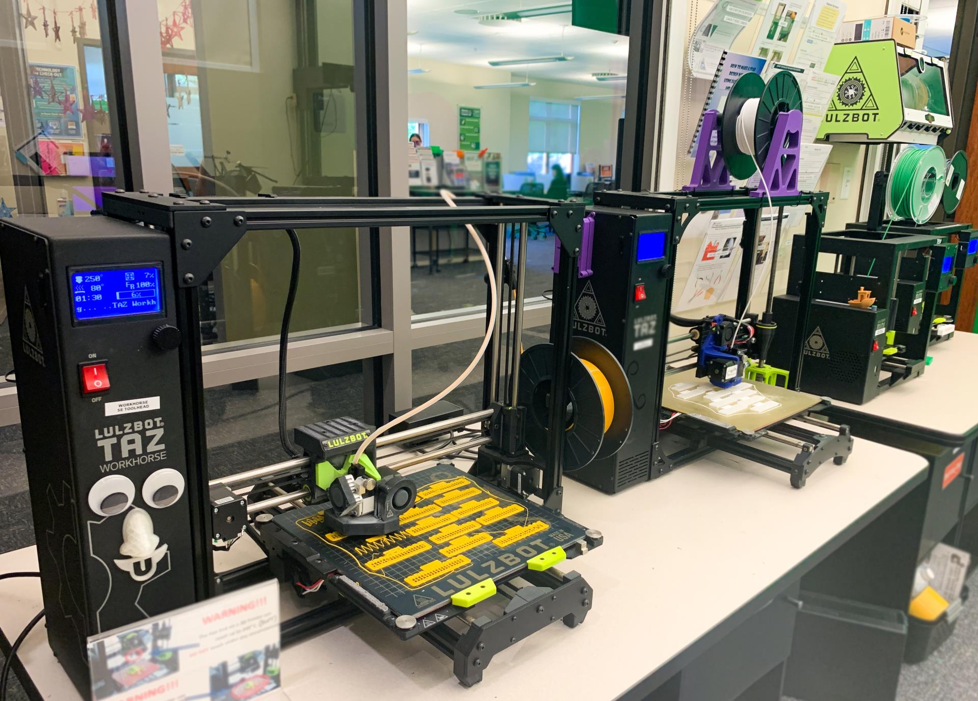 four Lulzbot branded 3D Printers printing in various colors of filament. Lulzbot Taz Workhorse in the foreground has been decorated with googly eyes and a 3d printed nose.