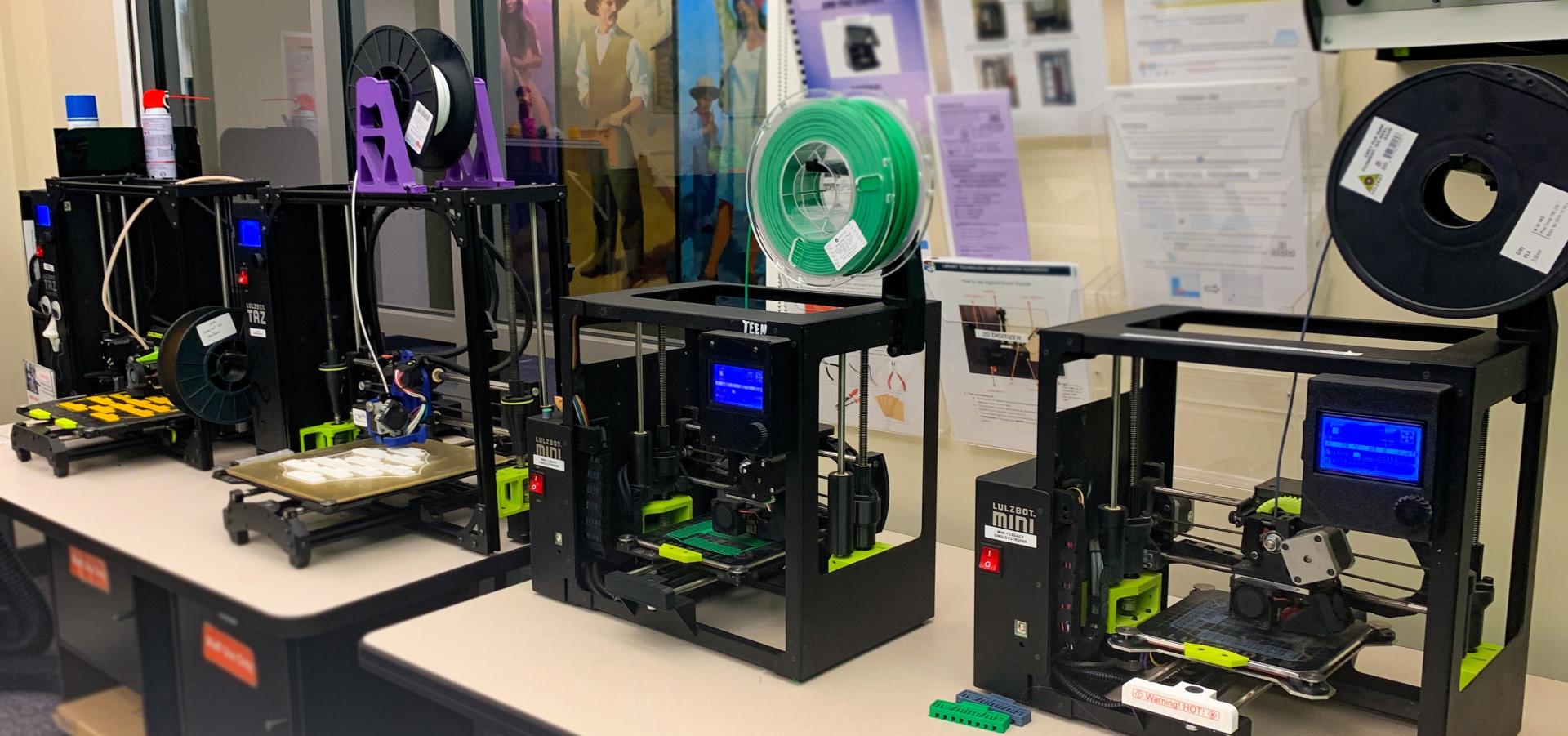 four lulzbot branded 3D printers printing in various colors of filament, informational guides and a mural can be seen in the background.